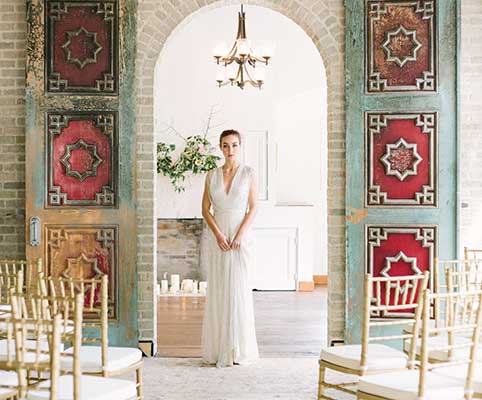 A bride in a white gown stands in the center of an arched doorway, framed by ornate red and gold doors. Rows of gold chairs and a chandelier are visible, suggesting a wedding ceremony setting.