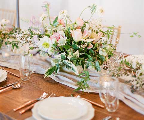 A rustic wooden table is set with white plates, glassware, silverware, and a flowing light gray runner. A lush floral centerpiece with pink and white flowers and greenery decorates the table.
