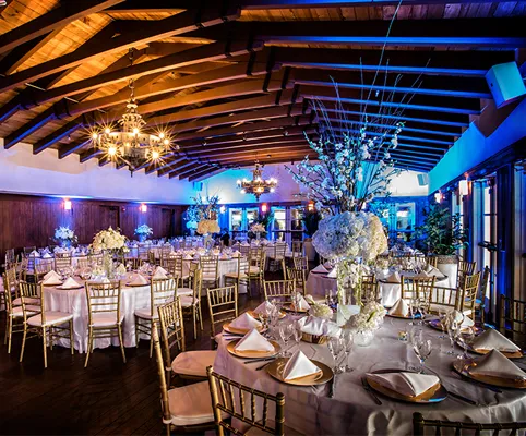 Elegant banquet hall set for an event with round tables covered in white linens, gold chairs, floral centerpieces, and soft lighting from chandeliers, creating a warm and inviting atmosphere.