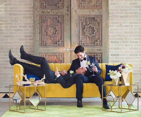 Two men in tuxedos relax on a yellow couch; one is lying with his head in the others lap. They are smiling and holding hands, surrounded by flowers and gold tables in a stylish, modern room with ornate doors.