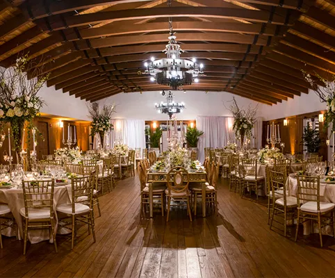 Elegant banquet hall with wooden floors and arched ceiling, set up for an event with round and rectangular tables adorned with white linens, gold chairs, floral centerpieces, and chandeliers providing warm lighting.