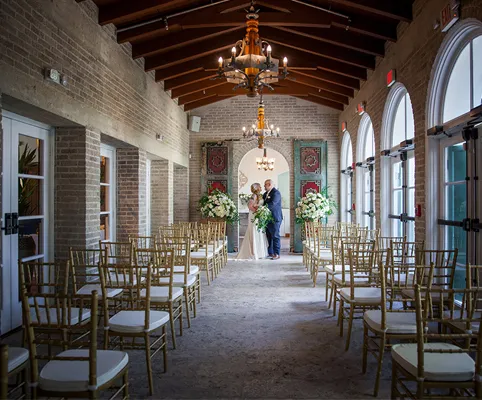 A bride and groom stand at the end of an aisle in a small, elegant indoor wedding venue with gold chairs and arched windows, decorated with white flowers and chandeliers.