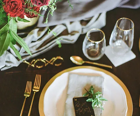 Elegant table setting with gold-rimmed plates and glasses, gold cutlery, a gray napkin, a small sprig of greenery, a black place card, a floral arrangement with red roses, and a gold “XOXO” decoration.