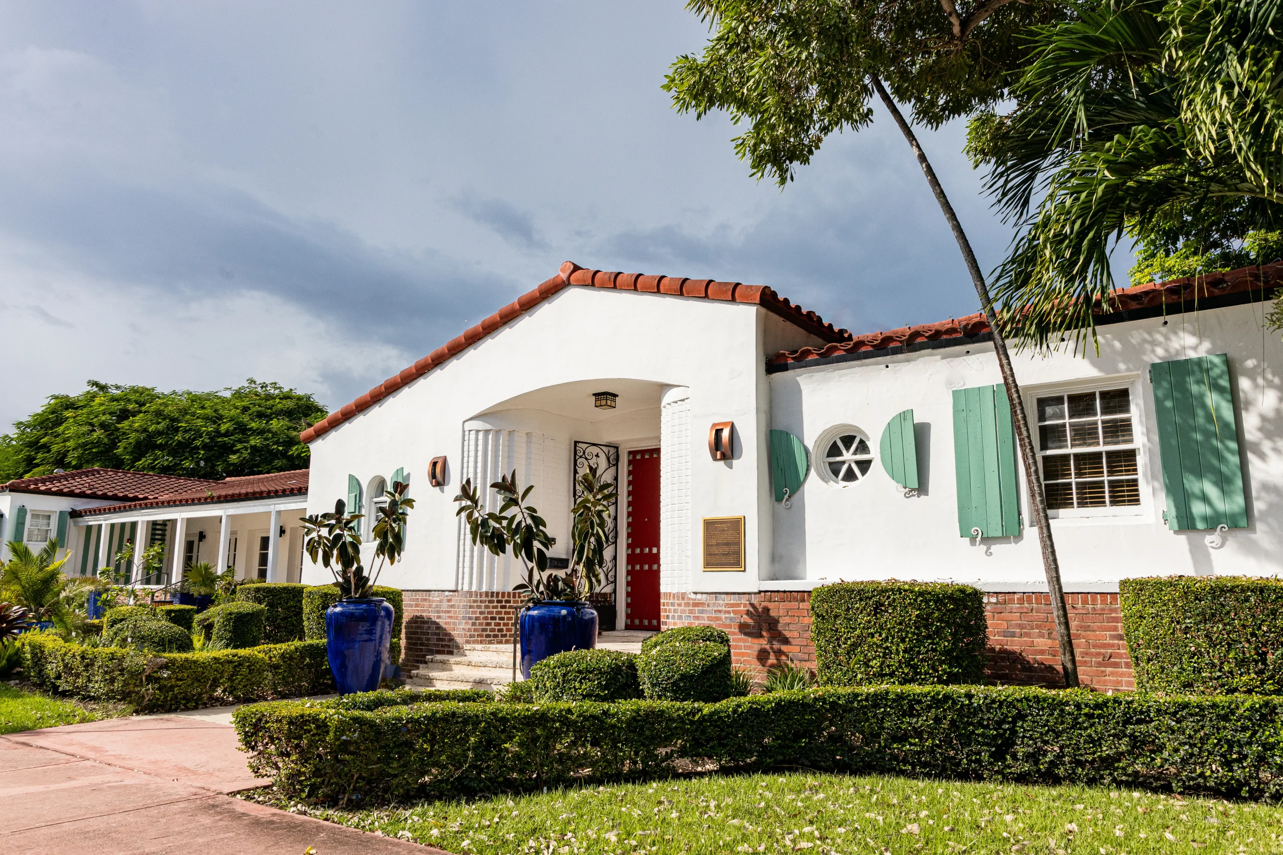 A white stucco building with a red tile roof, turquoise shutters, blue ceramic planters, and neatly trimmed hedges sits under a partly cloudy sky.