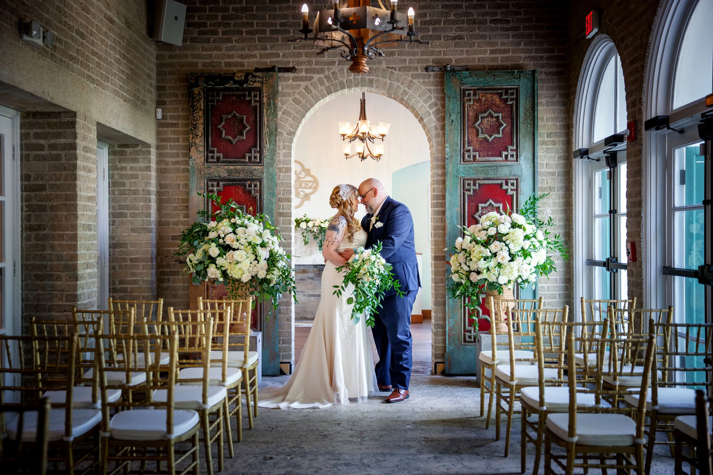 A bride and groom stand close together at the altar in an elegant indoor wedding venue, surrounded by gold chairs and large floral arrangements, with arched windows and rustic décor in the background.