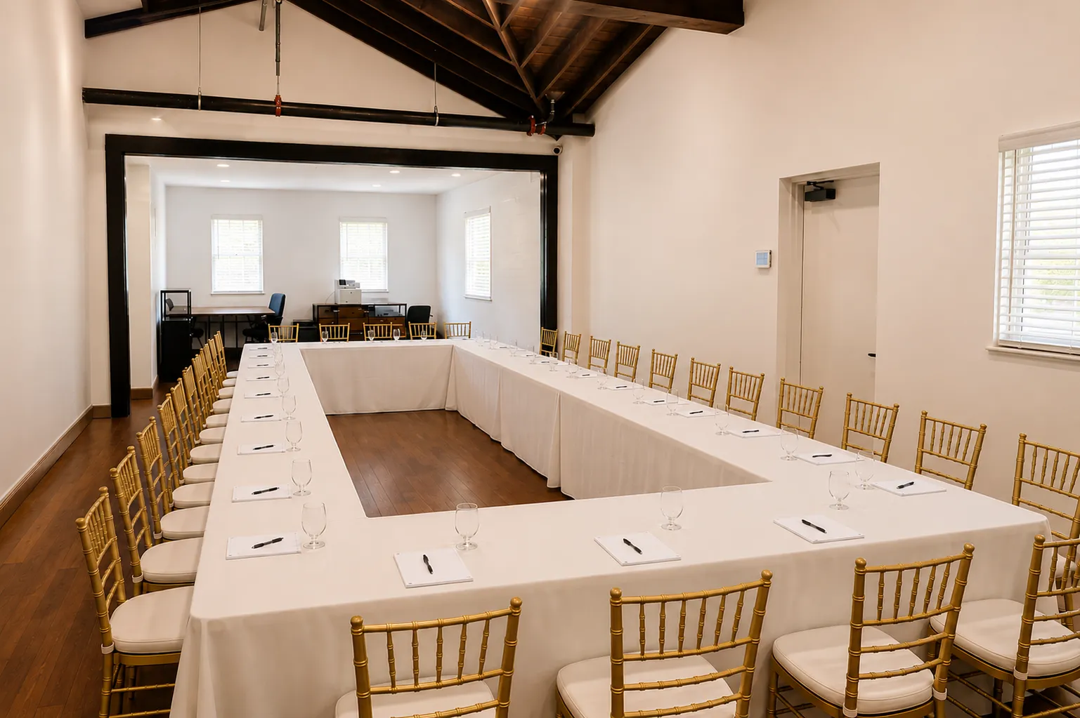 A conference room with a U-shaped table setup, covered in white tablecloths, surrounded by gold chairs. Each place setting has a notepad, pen, and water glass. Large windows let in natural light.