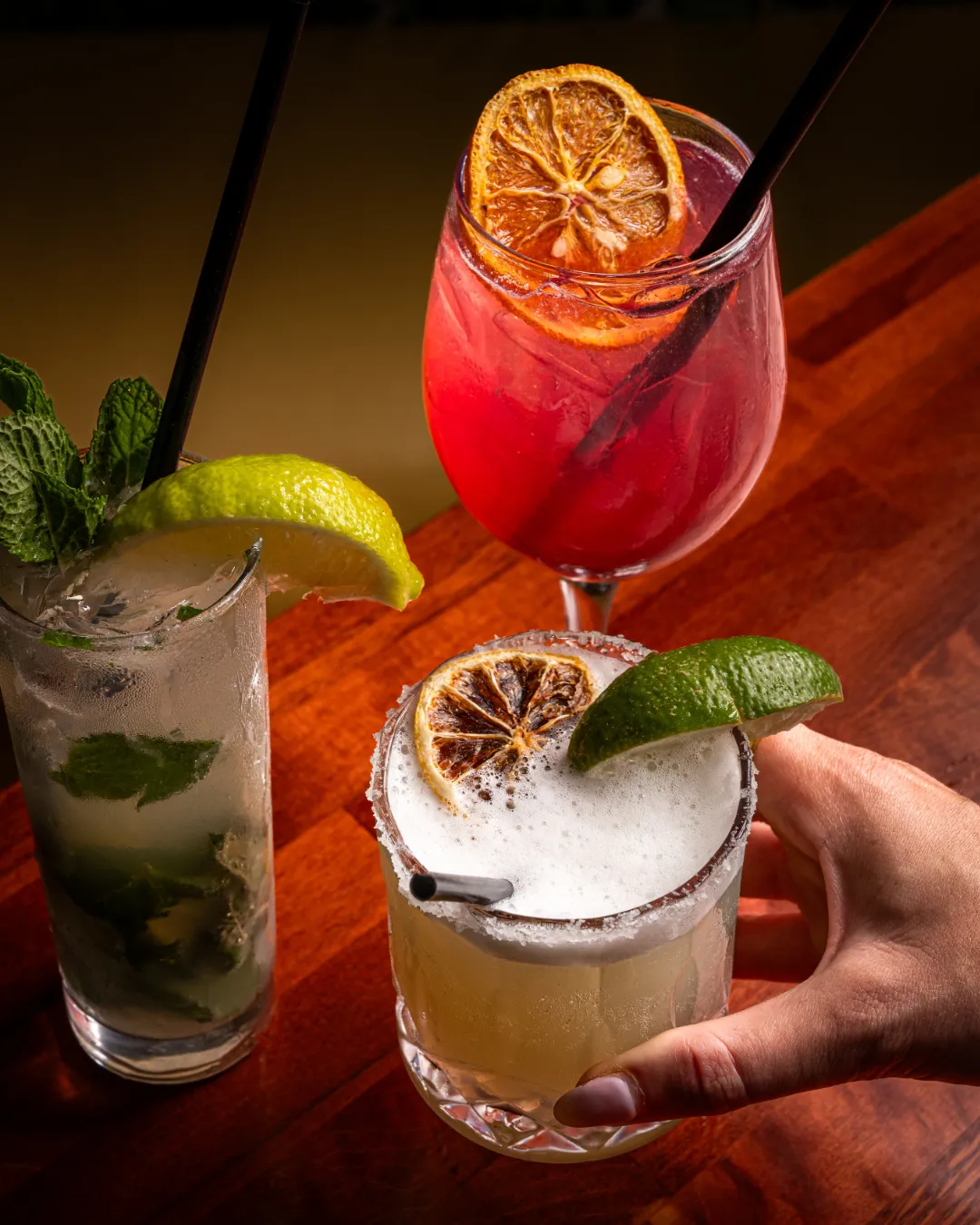 Three colorful cocktails on a wooden table: a mojito with mint and lime, a pink drink with dried orange slices, and a margarita with a lime wedge and dried orange, being held by a hand.