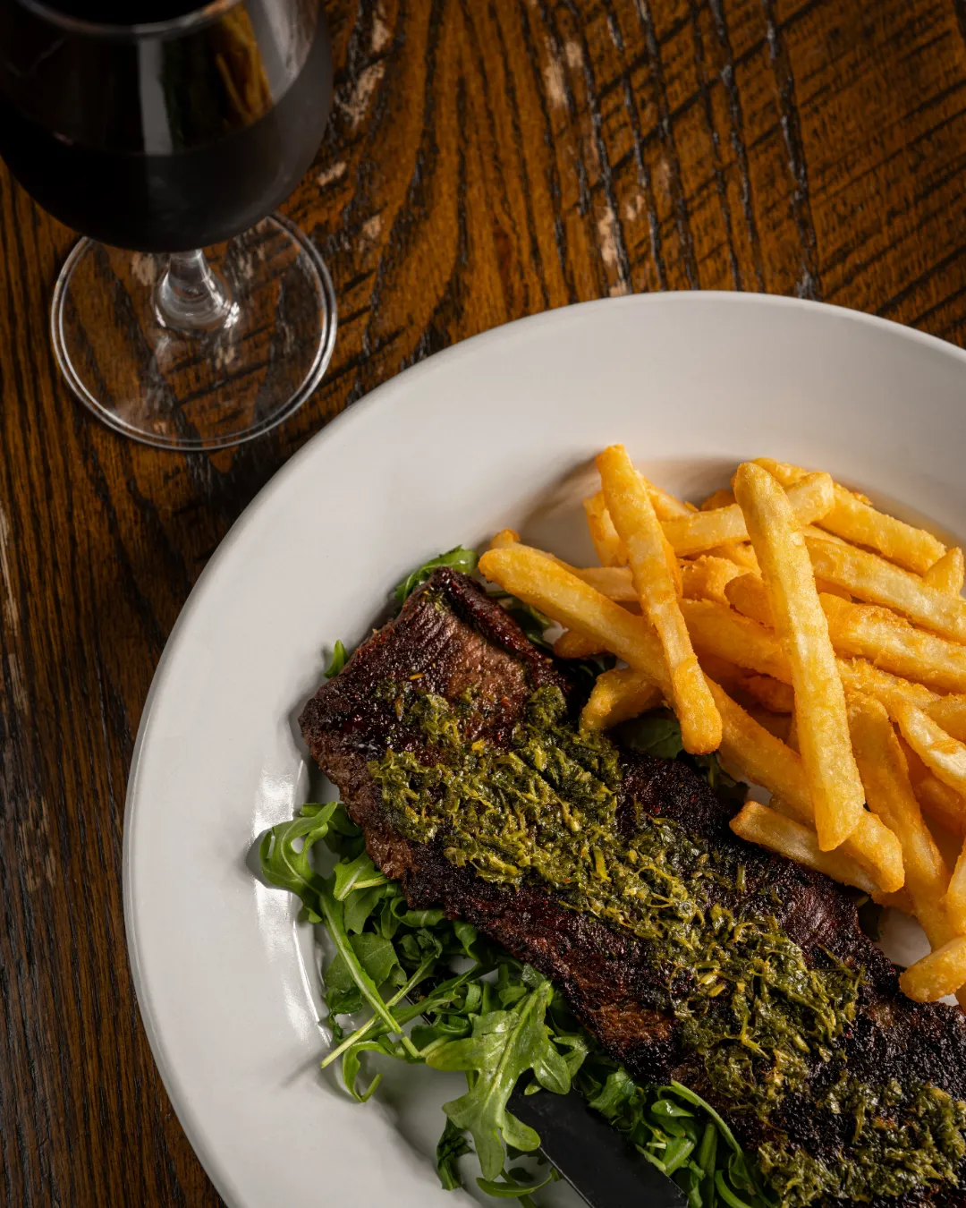 A plate with grilled steak topped with green herb sauce, served on a bed of arugula alongside a generous portion of golden French fries, sits on a wooden table next to a glass of red wine.