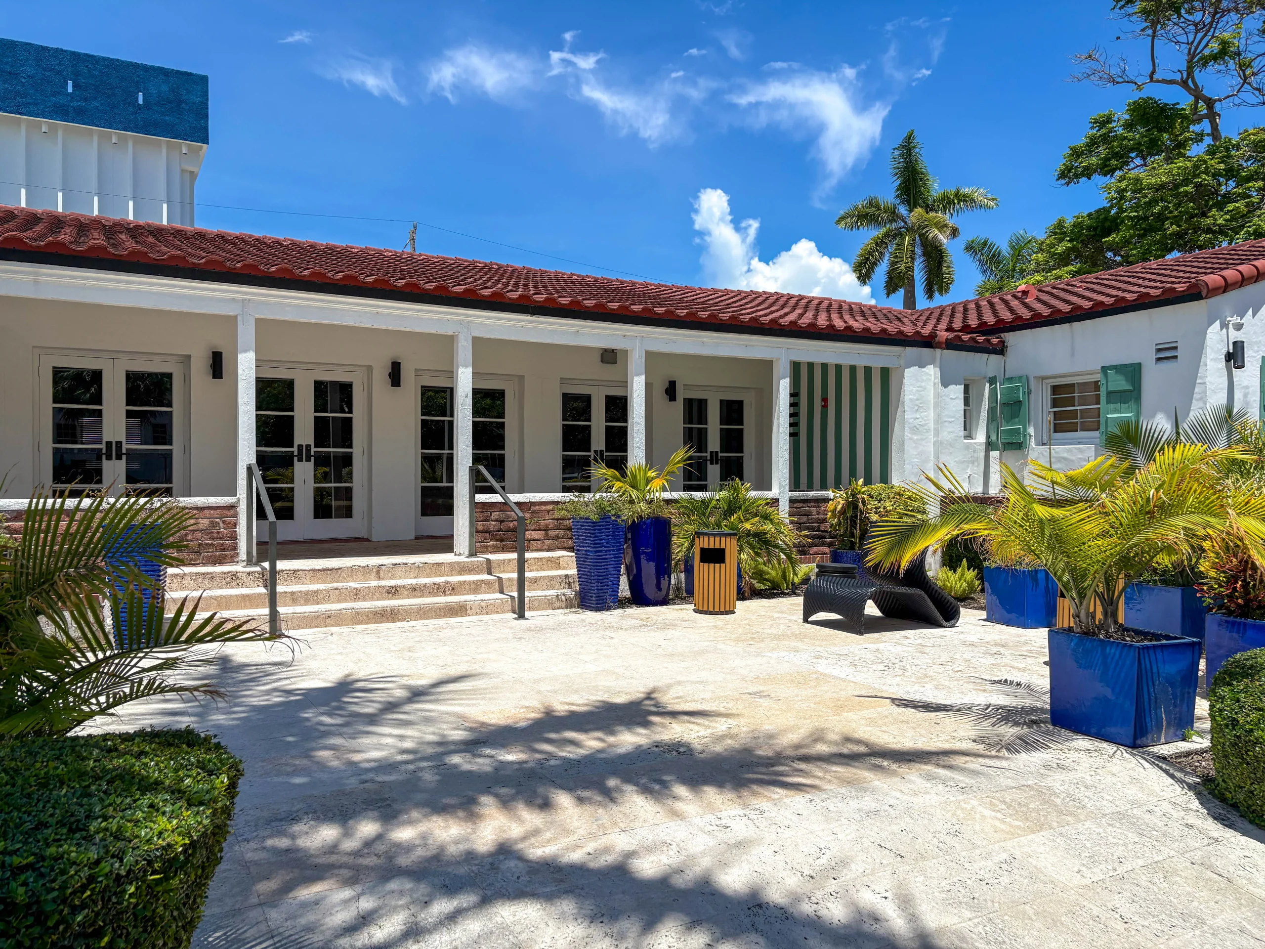 A sunny courtyard with tropical plants in blue planters, light stone pavement, and a white building with a red-tiled roof and multiple glass doors. Palm trees and blue sky are visible in the background.