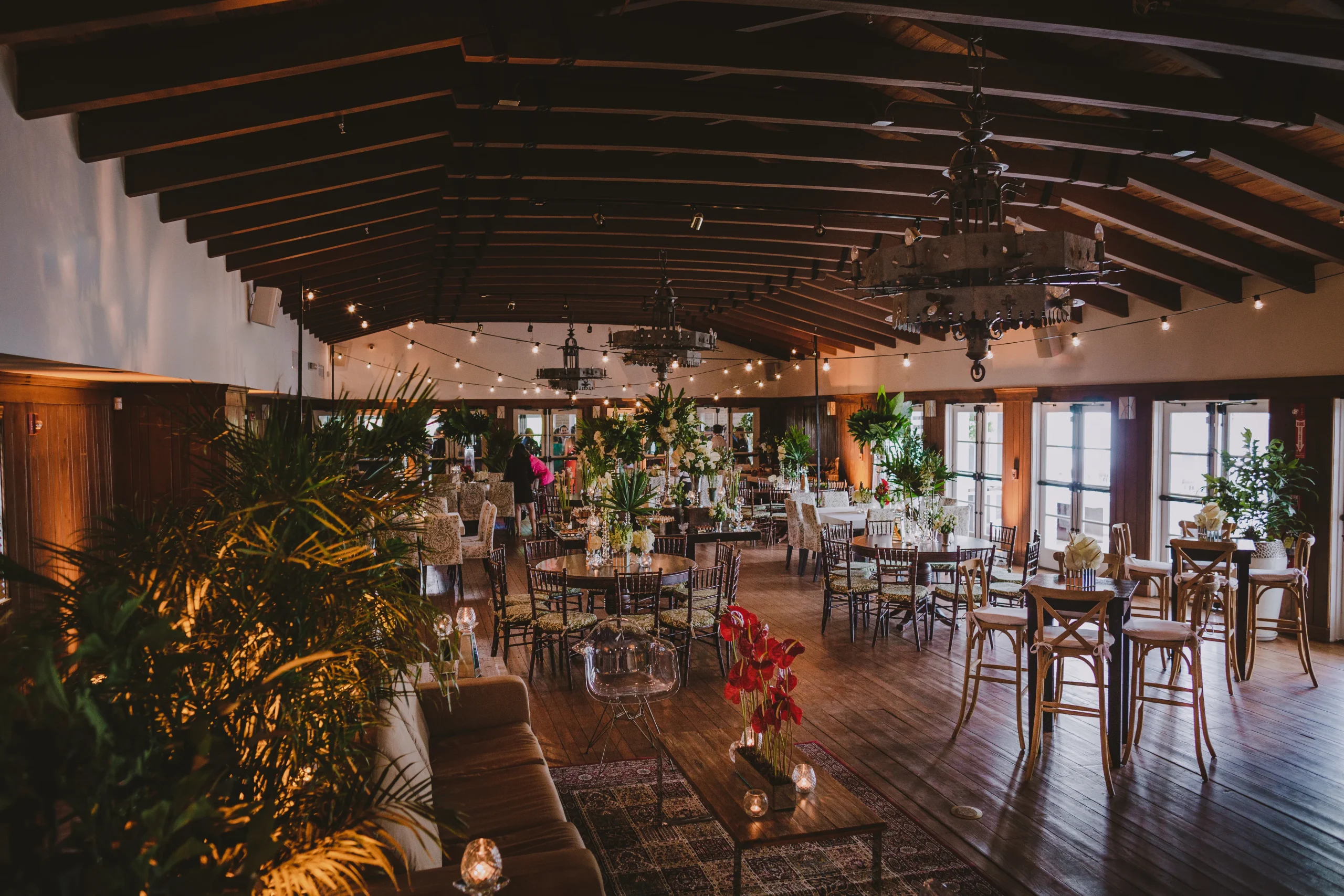 Elegant indoor event venue with wooden floors and ceiling, decorated with hanging chandeliers, lush green plants, round tables set for dining, and large windows allowing natural light to fill the spacious room.