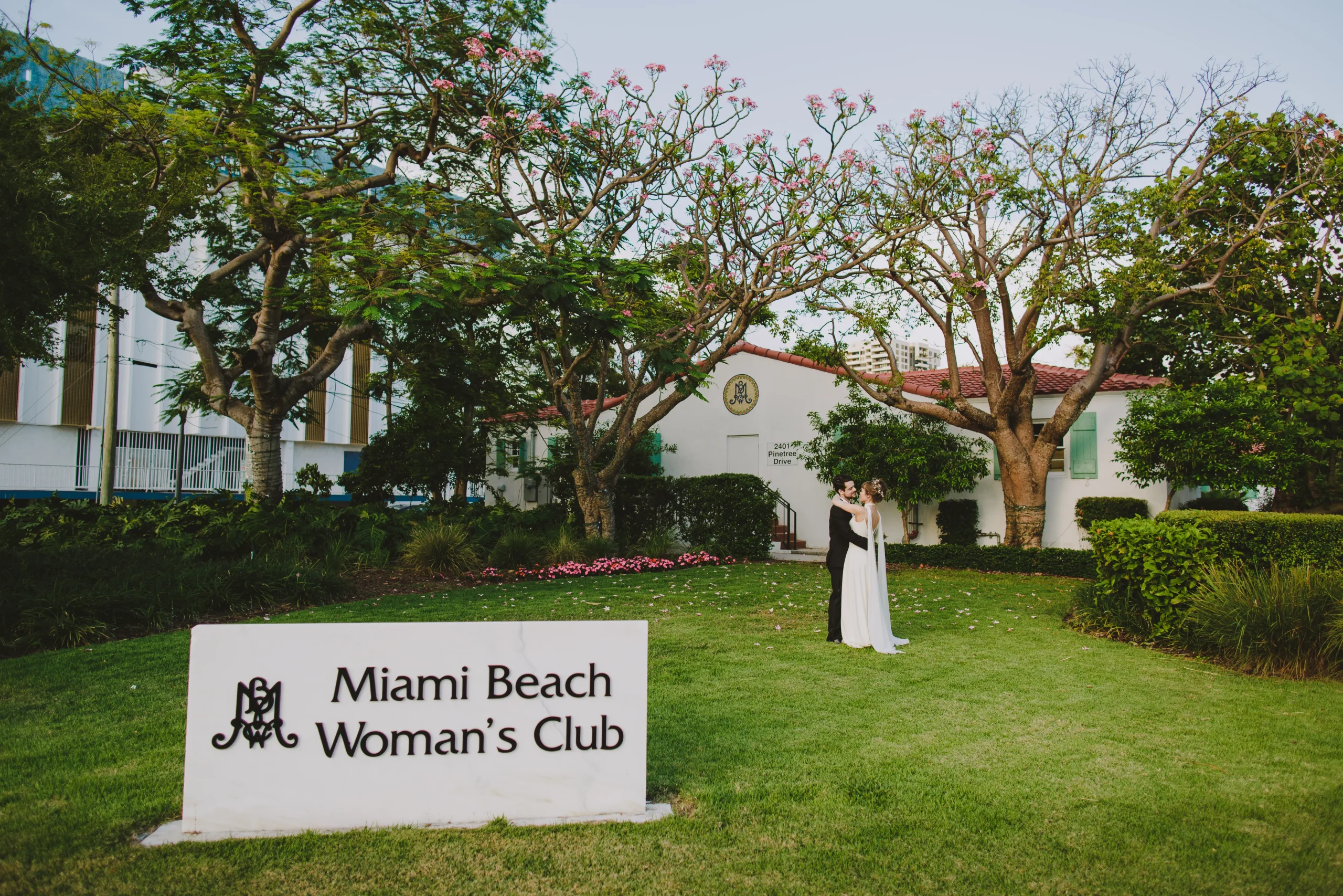 A couple dressed in formal attire stands on the lawn in front of the Miami Beach Woman’s Club building, surrounded by trees and greenery, with a large white sign in the foreground.