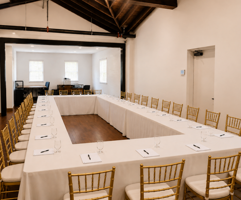 A conference room with a U-shaped table setup, covered in white tablecloths, surrounded by gold chairs. Each place has a notepad, pen, and water glass. The room has wooden floors, white walls, and large windows.