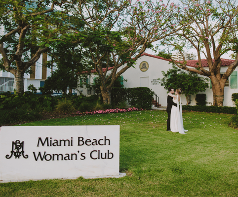 Two women in formal attire stand together on the lawn outside the Miami Beach Woman’s Club, surrounded by trees and greenery, with a large club sign visible in the foreground.