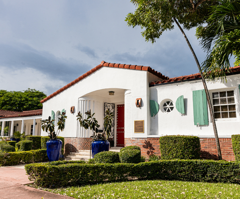 A white stucco building with a red tile roof, green shutters, and a bright red door. Two large blue planters with plants flank the entrance. Well-manicured bushes and trees surround the building on a sunny day.