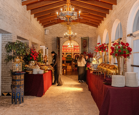 A lavish buffet setup in an elegant, narrow hall with arched windows, red tablecloths, gold chafing dishes, floral decorations, and guests serving themselves under warm chandelier lighting.