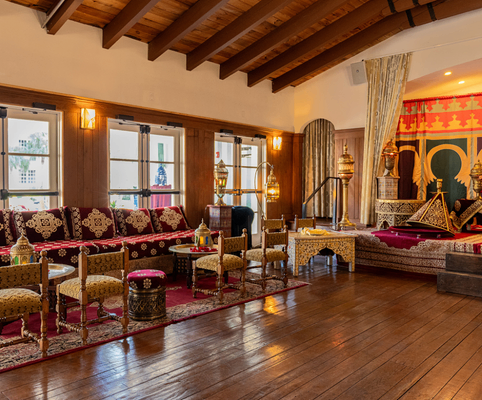 A luxurious living room with ornate gold and red furniture, intricate rugs, wooden floors, and a canopied bed, featuring vintage decor and large windows letting in natural light.