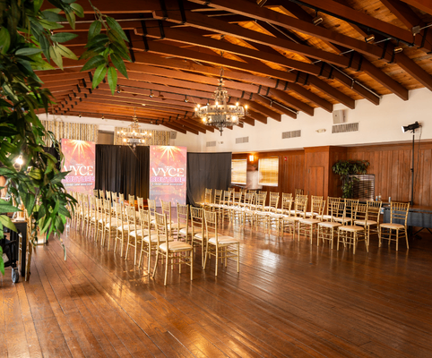 Spacious event hall with wooden floors and exposed beam ceiling, set up with gold chairs in rows facing a stage with colorful banners and chandeliers hanging overhead.