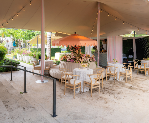 Outdoor seating area under a canopy with string lights, wooden chairs, round tables covered with white tablecloths, and a central pink umbrella over an arrangement of flowers. Trees and plants surround the space.