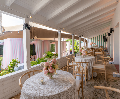 Outdoor patio with wooden chairs and round tables covered in white patterned tablecloths, each decorated with a pink floral centerpiece. The area is shaded by a white roof and overlooks lush greenery and pastel cabanas.