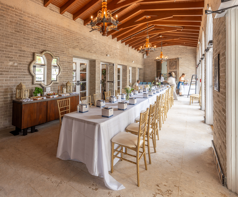 A long dining table with a white tablecloth and gold chairs is set for a meal in a bright, elegant room with brick walls, high wooden ceiling, chandeliers, and large windows letting in natural light.