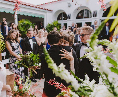 A bride embraces another person tightly at an outdoor wedding ceremony, surrounded by guests in formal attire and lush floral decorations, with onlookers smiling in the background.