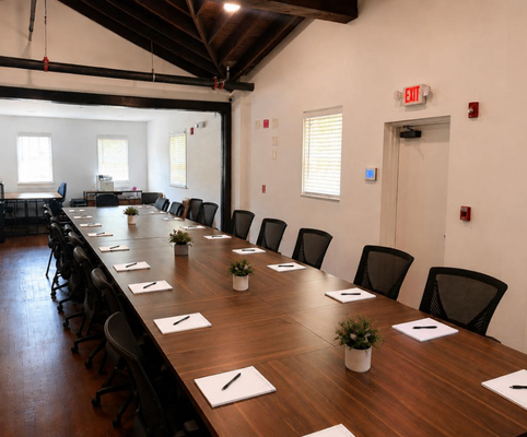 A large conference room with a long wooden table surrounded by black chairs, each place set with a notepad, pen, and a small potted plant. The room has white walls and several windows letting in natural light.