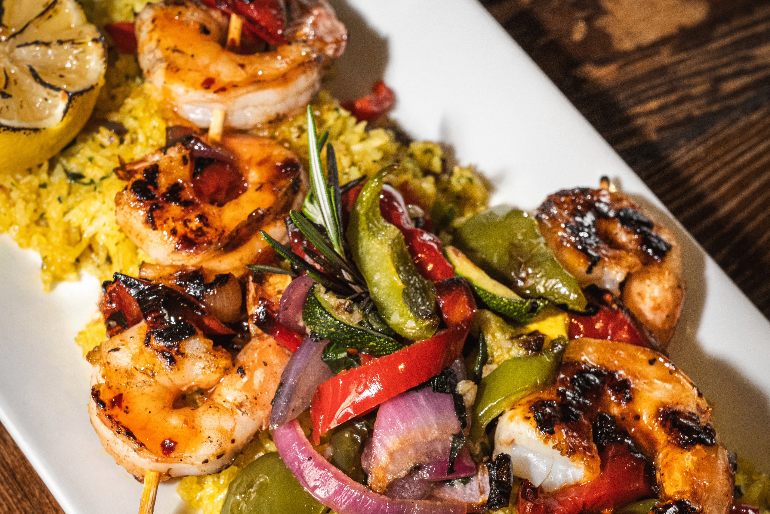 Grilled shrimp skewers with bell peppers, red onions, and zucchini served over yellow rice, garnished with a grilled lemon wedge and fresh rosemary on a white plate.