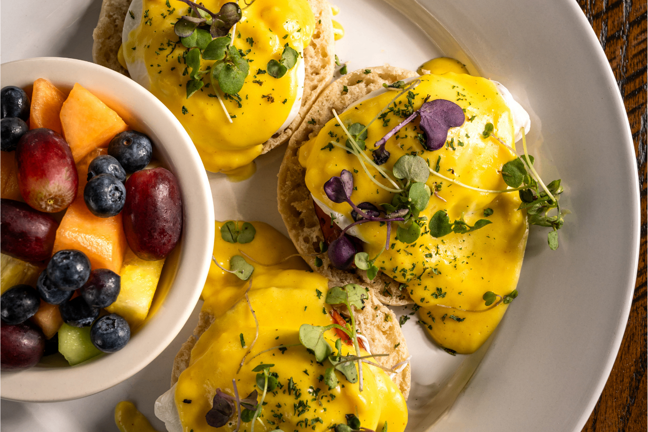 A plate with three eggs benedict topped with yellow hollandaise sauce and microgreens, served next to a bowl of mixed fruit including grapes, blueberries, melon, and pineapple.
