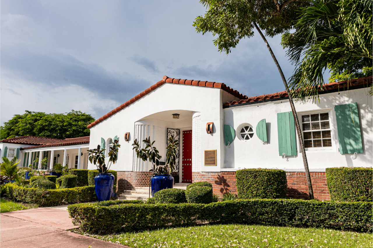 A white stucco house with a red tile roof, green shutters, and blue ceramic pots near the entrance, surrounded by manicured bushes and palm trees under a partly cloudy sky.